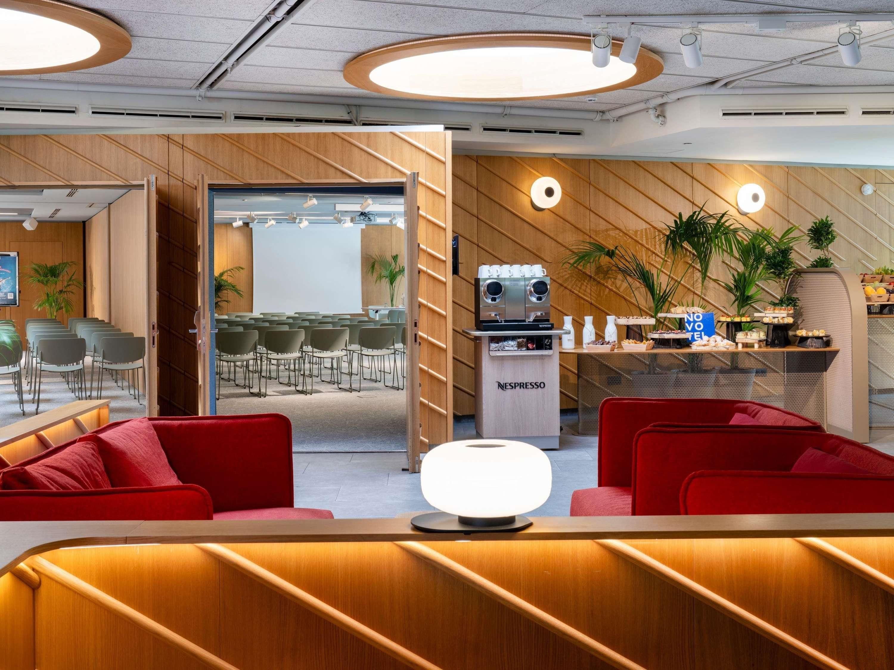 Een moderne lounge met rode banken, een koffiehoek en een zichtbare vergaderruimte door een open deur.