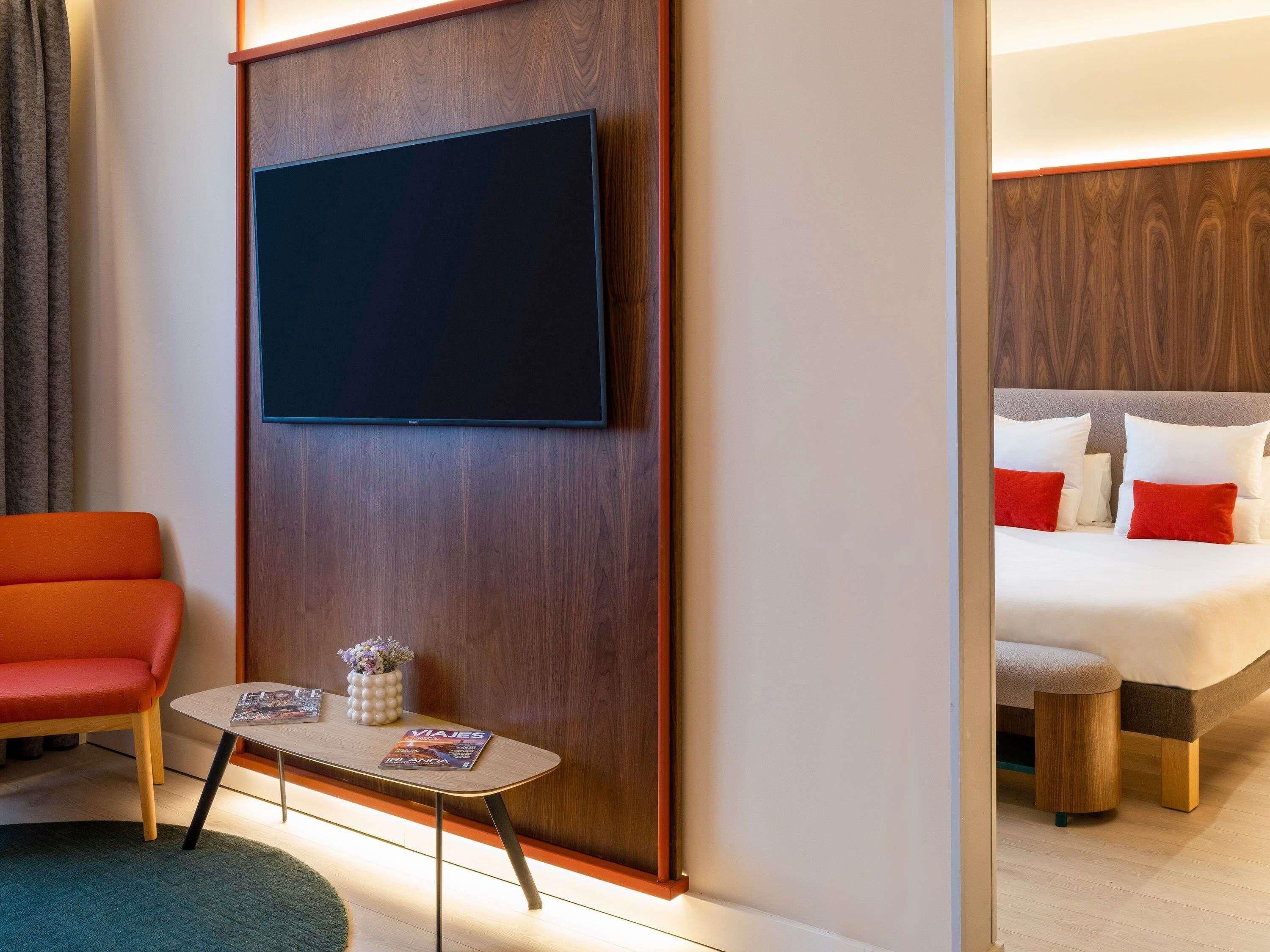 A hotel room with a bed, a TV mounted on a wooden panel, a small table with a magazine, a red chair, and a wooden nightstand.