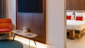 Habitación de hotel con una cama, un televisor montado en un panel de madera, una mesita con una revista, una silla roja y una mesita de noche de madera.