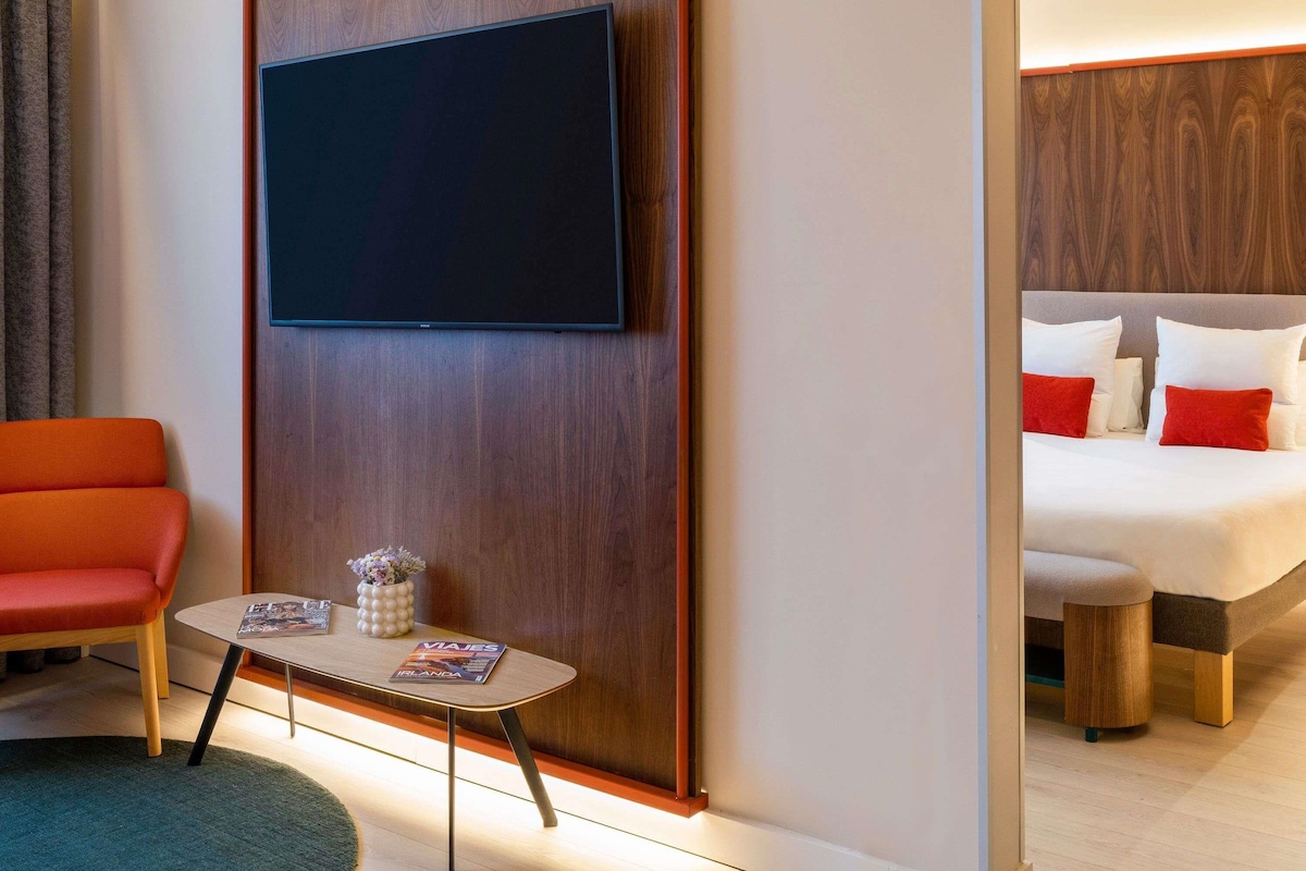 Habitación de hotel con una cama, un televisor montado en un panel de madera, una mesita con una revista, una silla roja y una mesita de noche de madera.