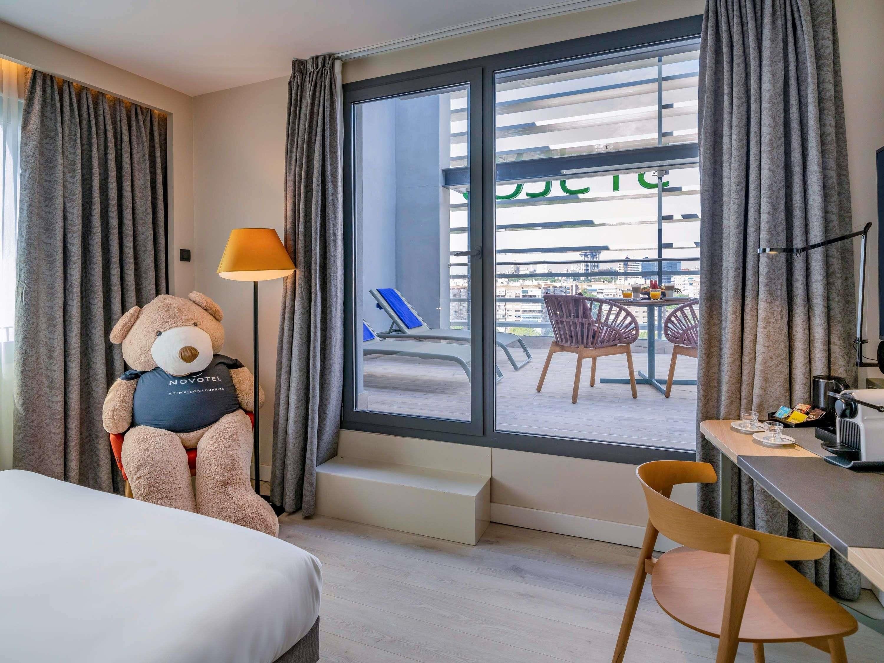 Habitación de hotel con un ventanal grande, una cama, un escritorio con silla, una mesa con silla y un peluche.