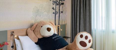 A bed with a large teddy bear, a wall with a tree mural, a bedside lamp, and a window with curtains.