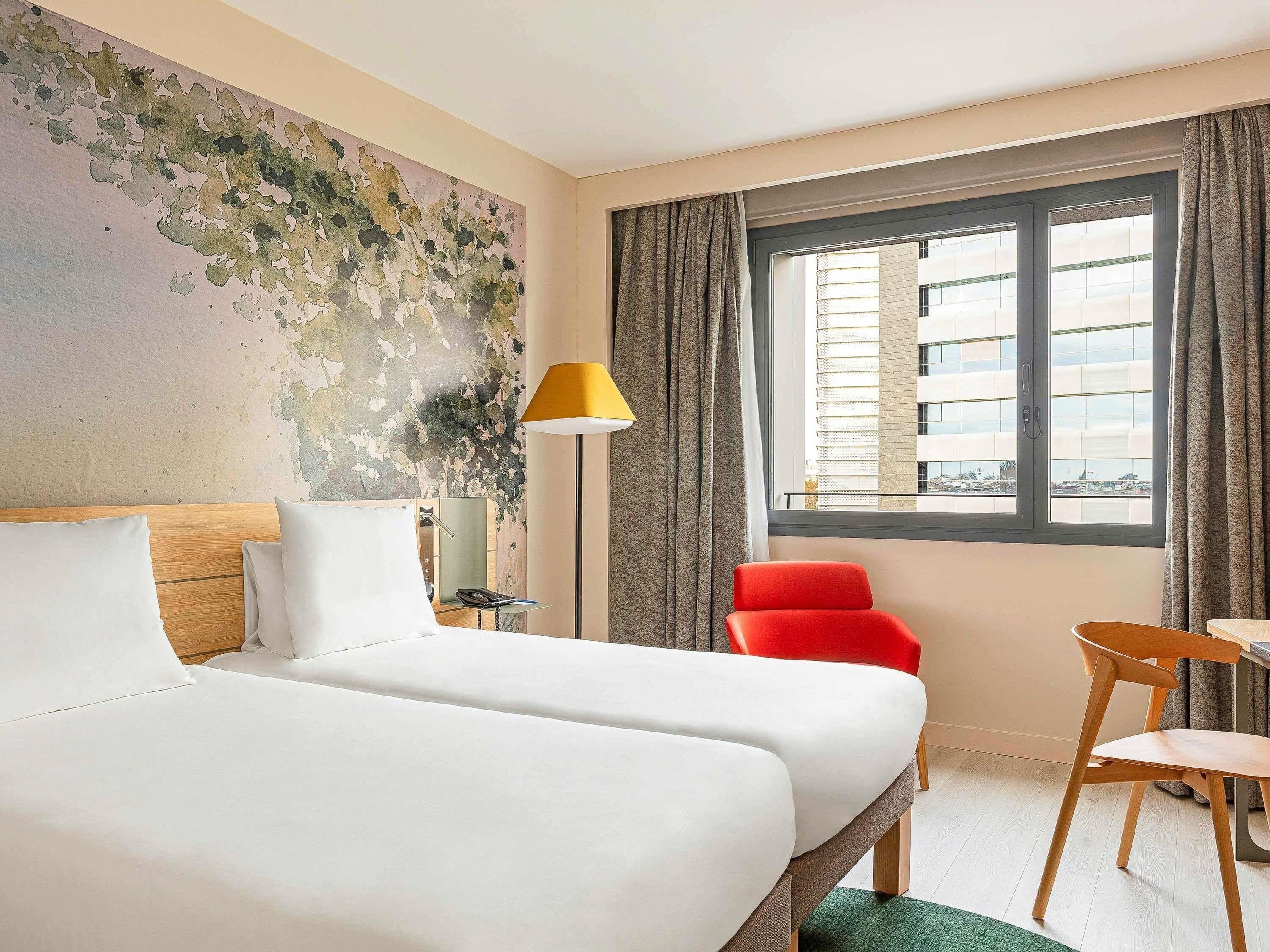 A hotel room with two beds, a red chair, a desk, a lamp, and a window with a city view.