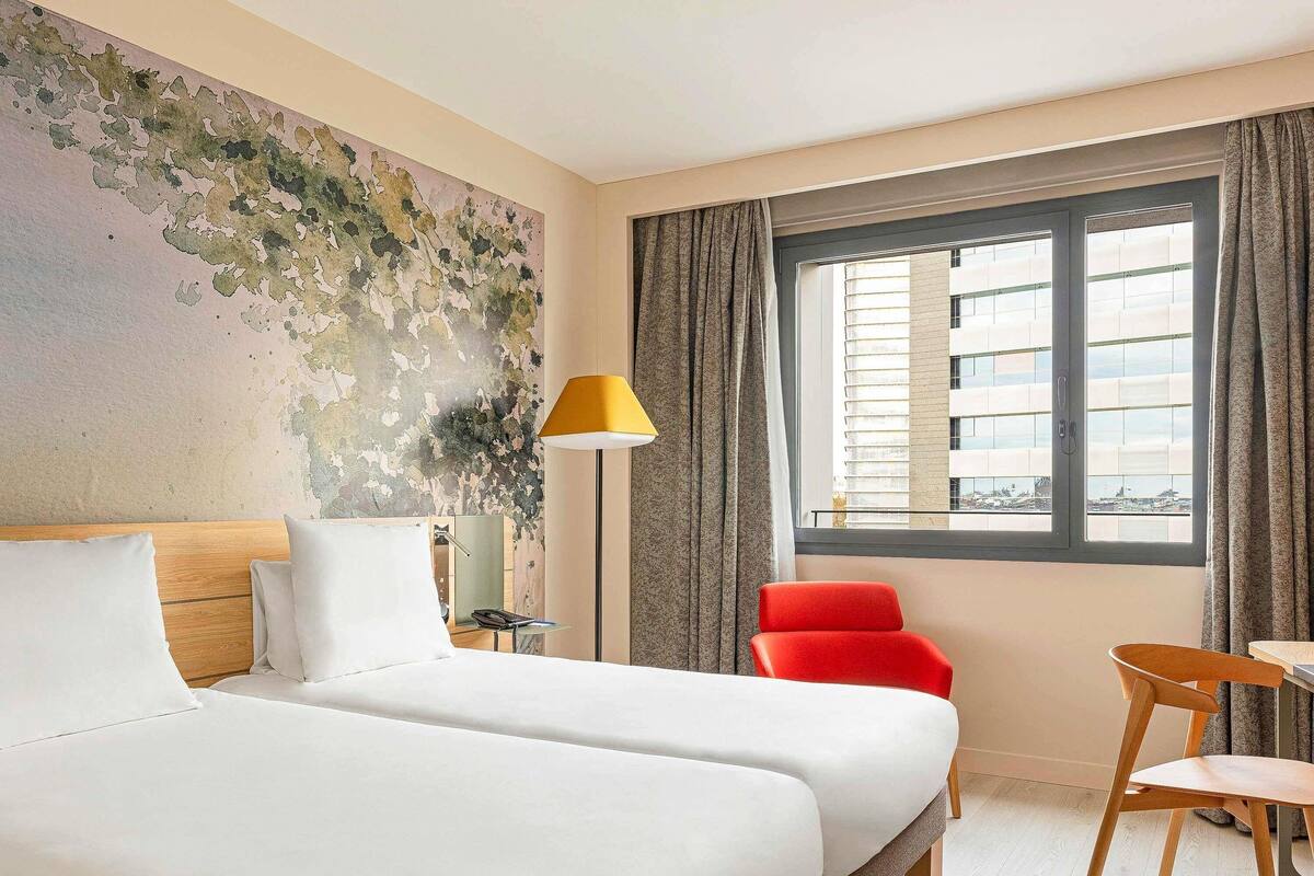 A hotel room with two beds, a red chair, a desk, a lamp, and a window with a city view.