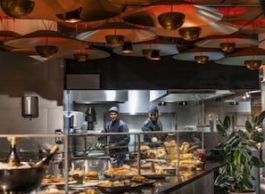A modern kitchen with stainless steel appliances, a buffet counter with various dishes, and overhead lighting. - Novotel Madrid City Las Ventas (Madrid)