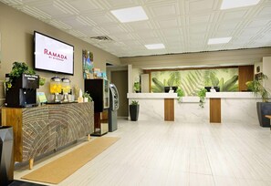 Un área de recepción con diseño moderno, que incluye un mostrador de recepción, una pantalla de televisión que muestra 'RAMADA BY WYNDHAM' y una pared con un mural tropical.