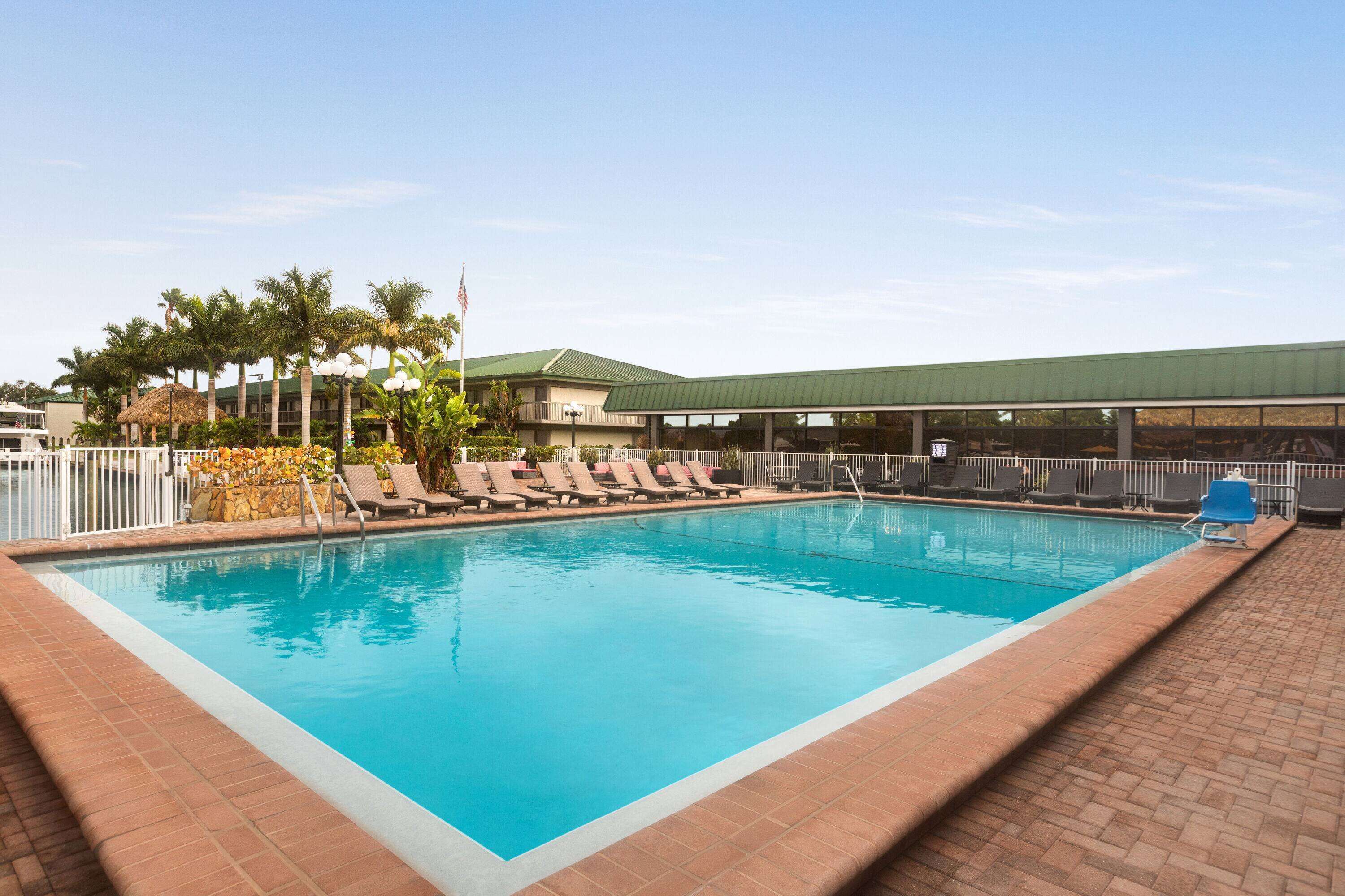 A rectangular swimming pool with clear blue water, surrounded by a tiled deck. There are lounge chairs along the pool's edge, and a building with a green roof in the background.