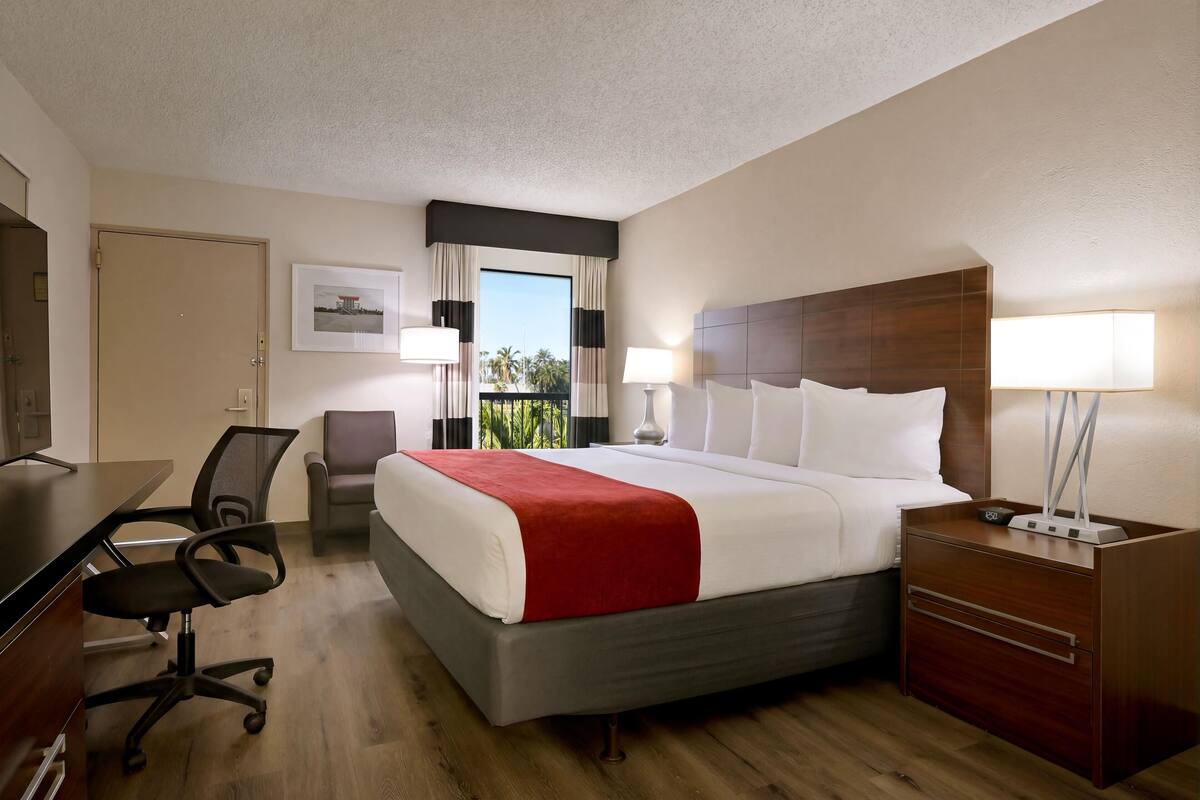 Une chambre d’hôtel avec un lit, un bureau, une chaise et une vue sur des palmiers.