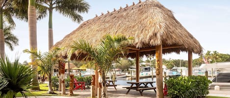 A thatched-roof pavilion with picnic tables, surrounded by palm trees and a marina with boats.