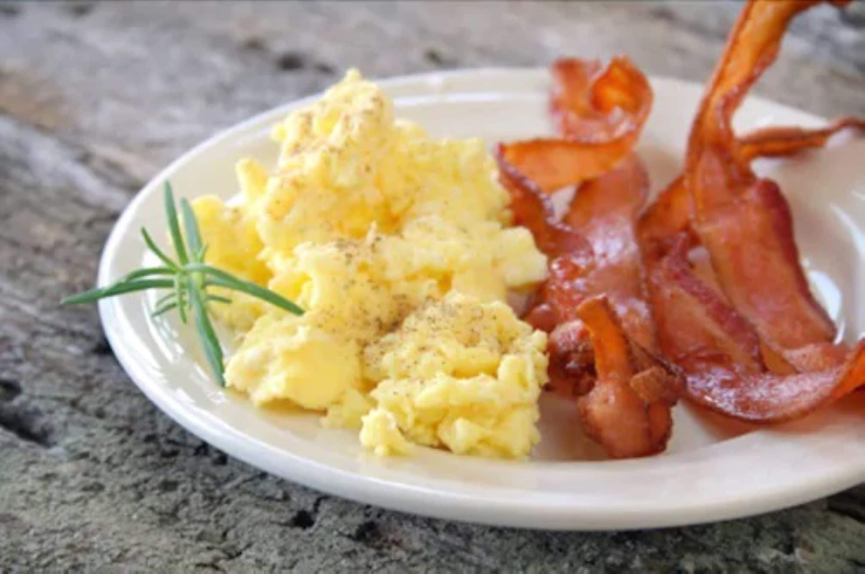 A plate with scrambled eggs, bacon, and a sprig of rosemary.