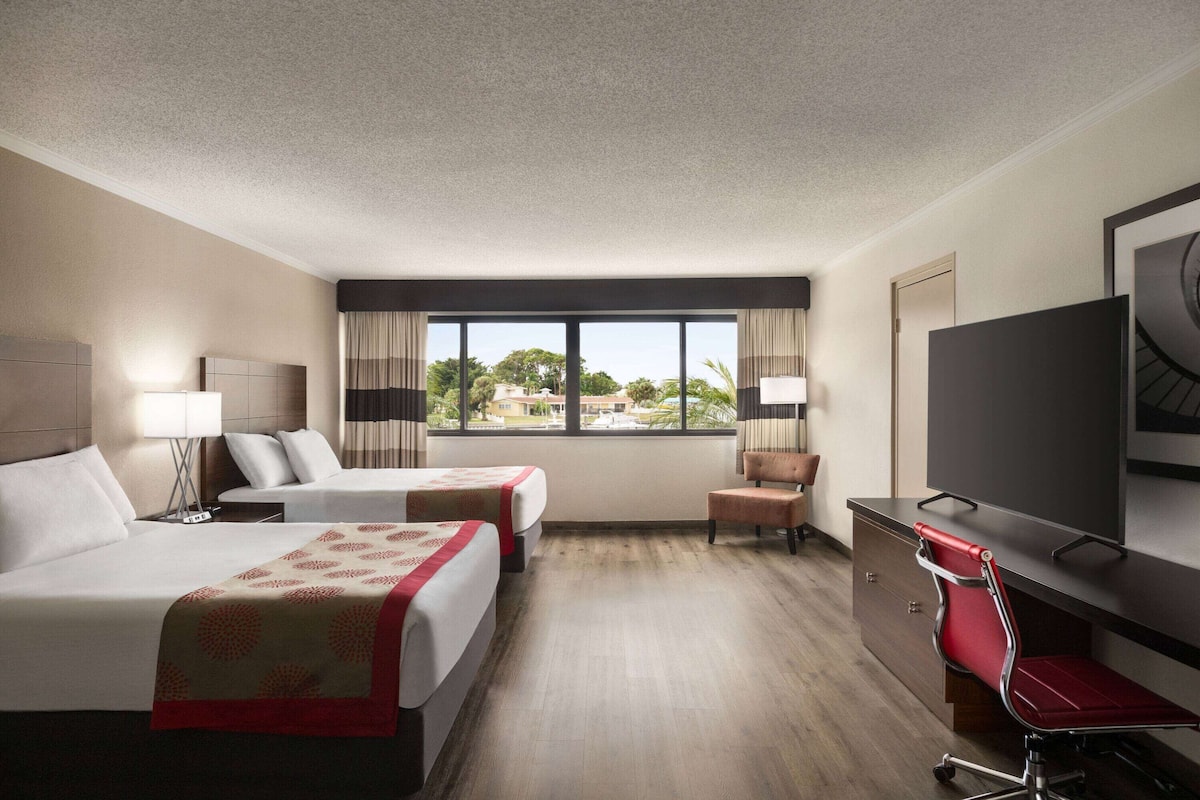 Une chambre d’hôtel avec deux lits, un bureau, une chaise, une télévision et une fenêtre donnant sur des immeubles et des arbres.
