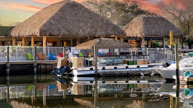 Una zona ribereña con construcciones de techo de paja, un muelle con barcos y un letrero que dice 'Low Tide Tours'.