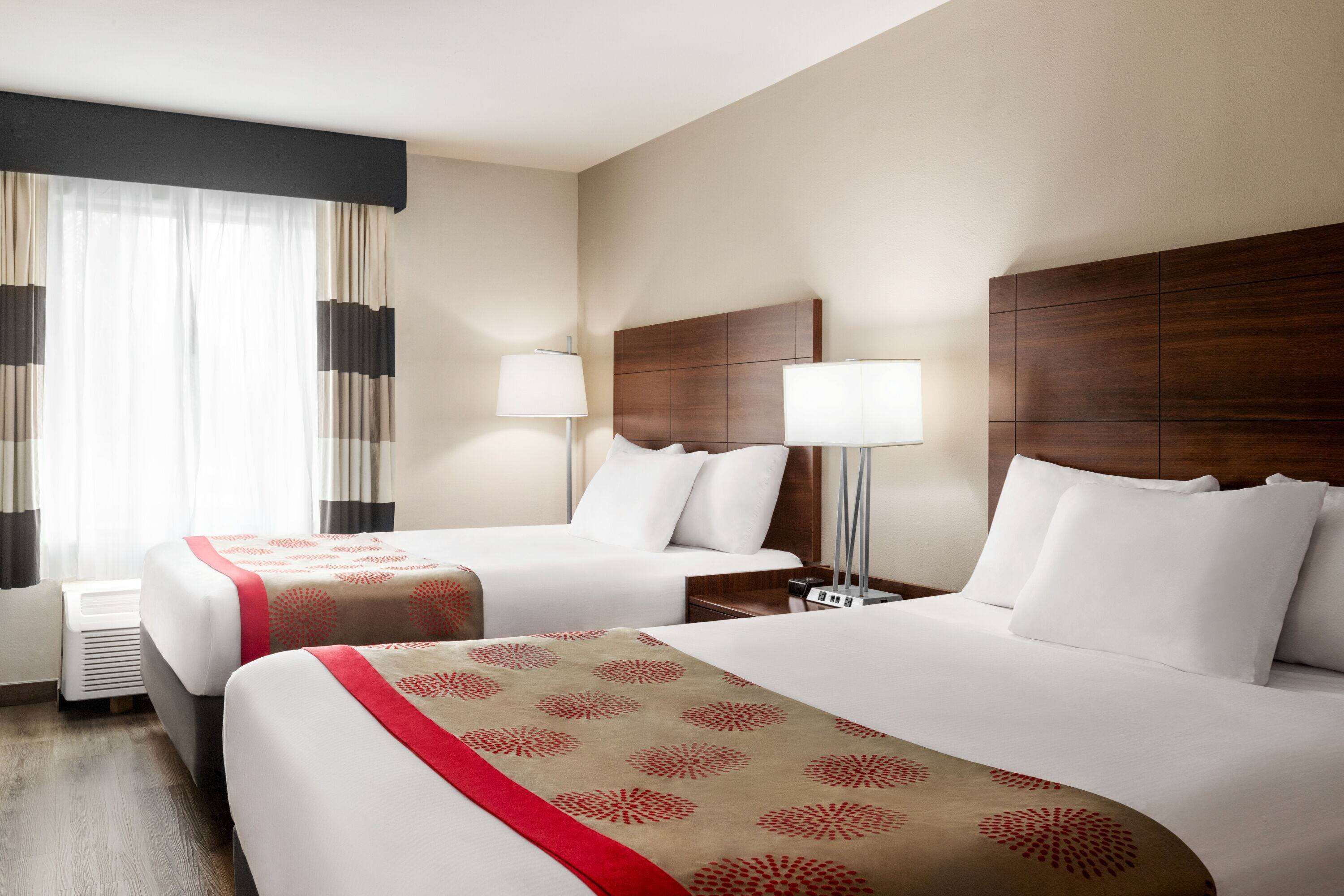 A hotel room with two beds, a wooden headboard, bedside lamps, and a window with sheer curtains.