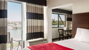 Habitación de hotel con cama, escritorio y silla. Amplia ventana con cortinas, y vista a un cuerpo de agua y vegetación.
