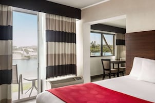 Ein Hotelzimmer mit Bett, Schreibtisch und Stuhl. Ein großes Fenster mit Vorhängen bietet Blick auf ein Gewässer und viel Grün.