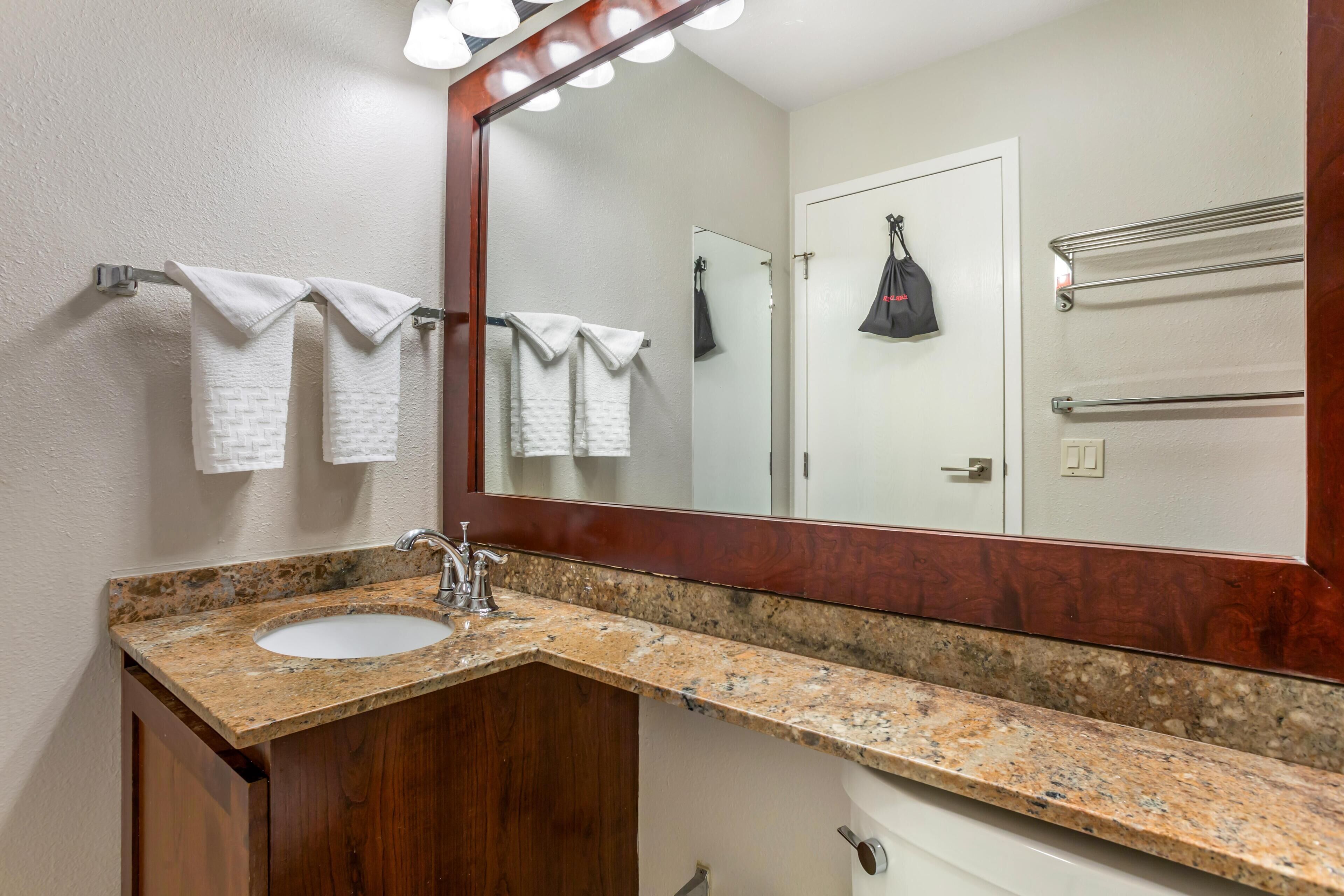 A bathroom with a granite countertop sink, a wooden cabinet, a large mirror, and a towel rack with white towels.