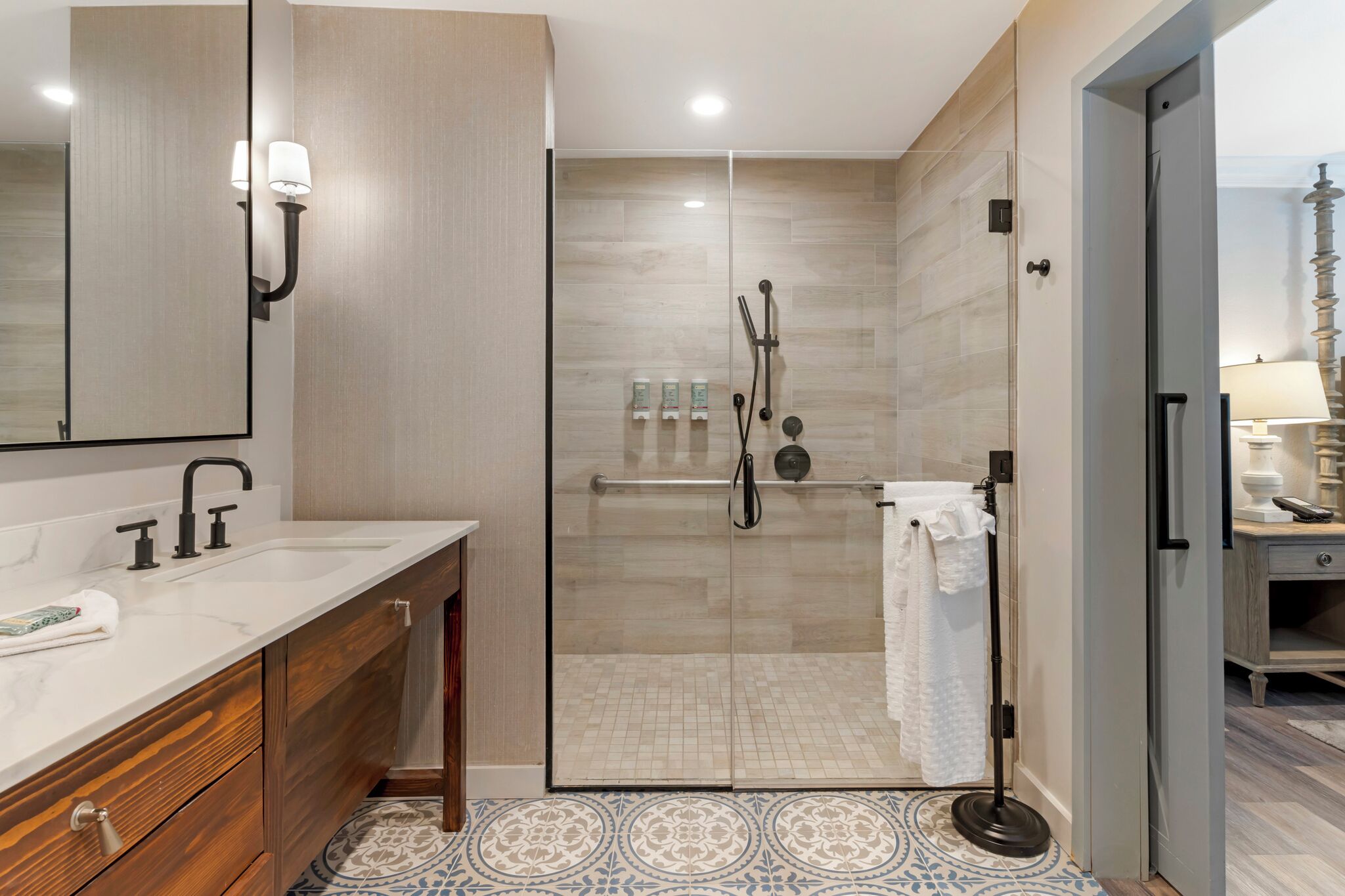A modern bathroom with a glass-enclosed shower, a wooden vanity with a marble countertop, and a patterned floor.