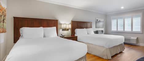 A hotel room with two beds, wooden flooring, a nightstand with a lamp, and a window with blinds.