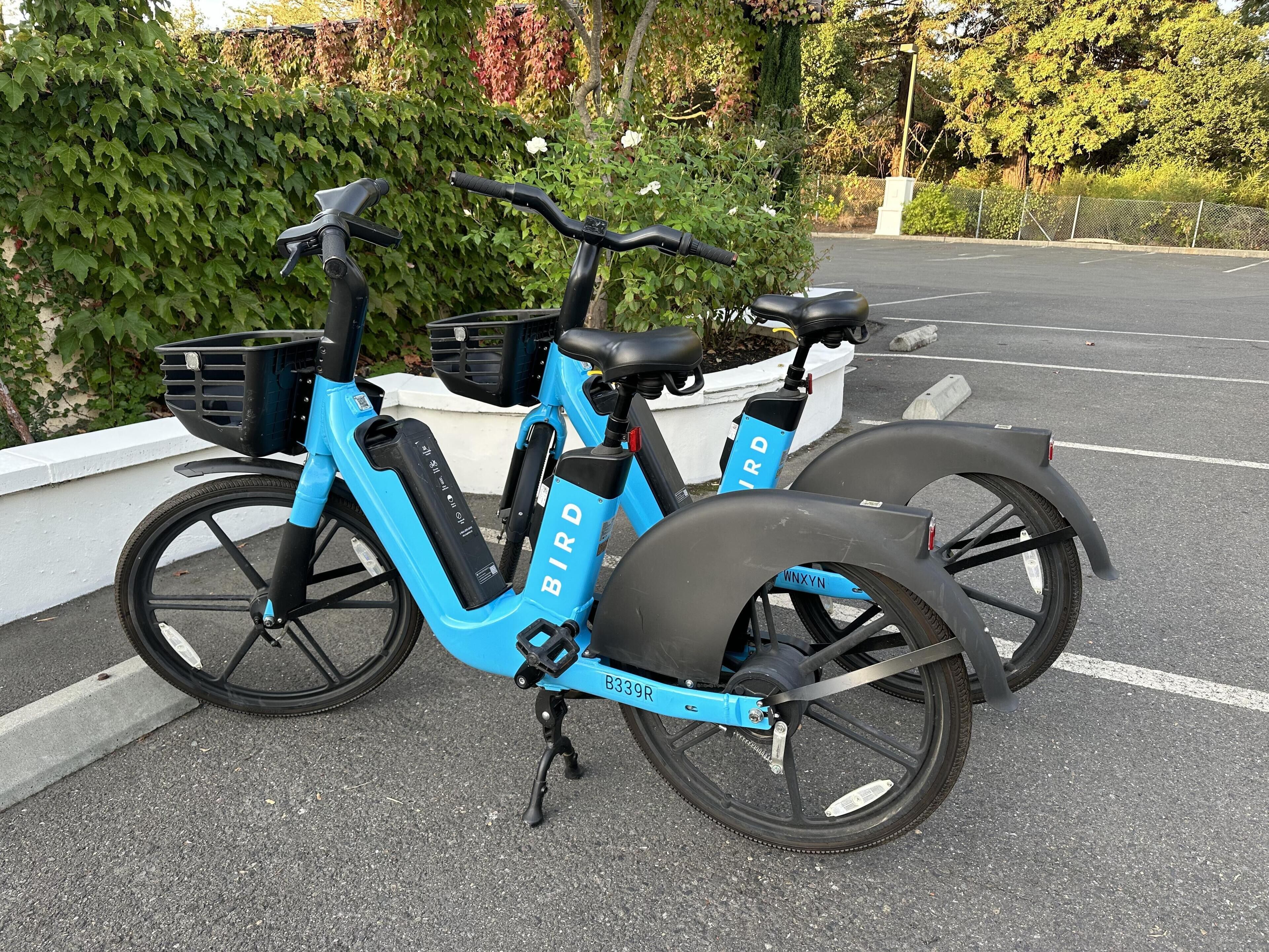 A blue electric bicycle with a basket and a rear rack parked in a parking lot.