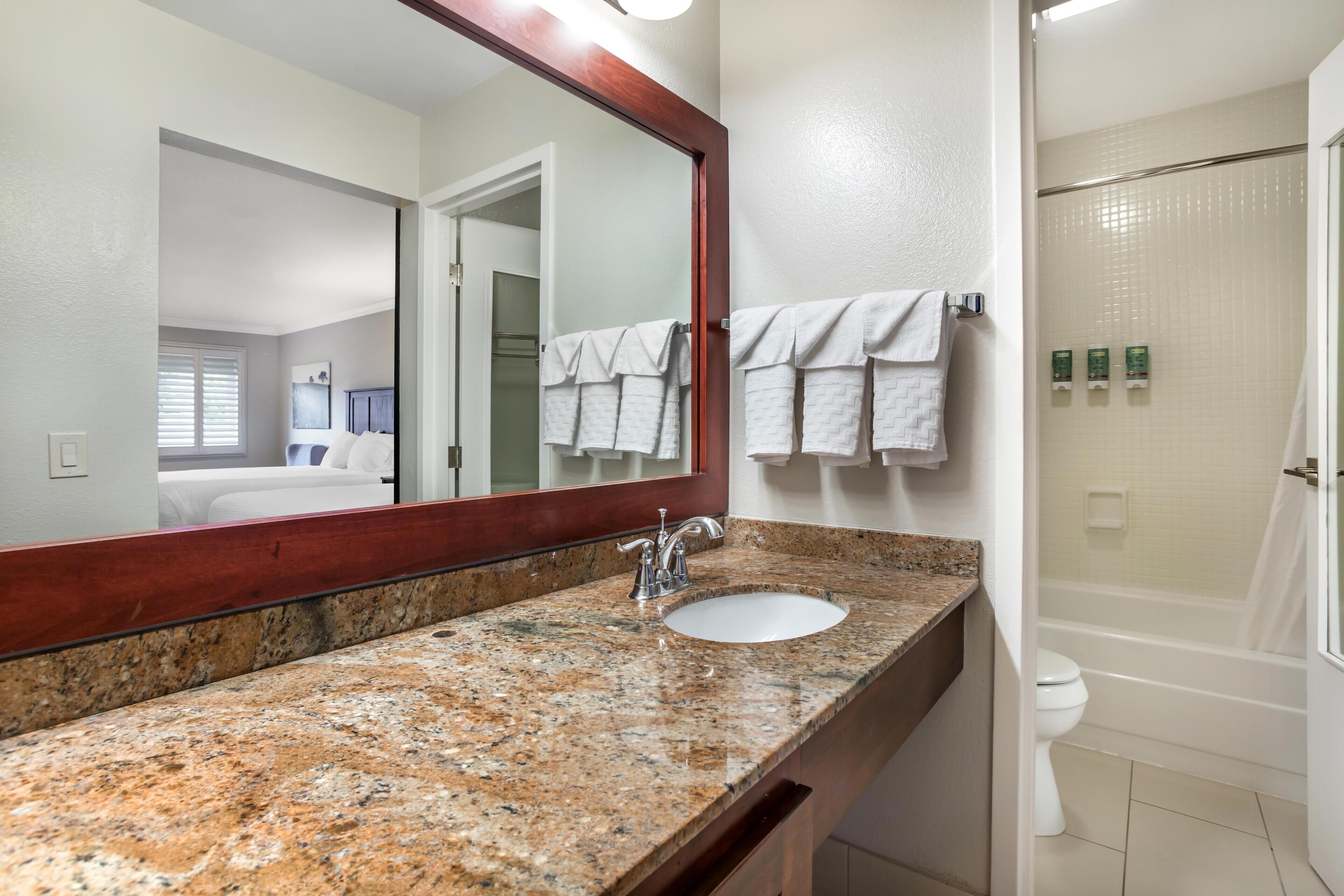 A bathroom with a granite countertop sink, a large mirror, a shower, and a toilet.