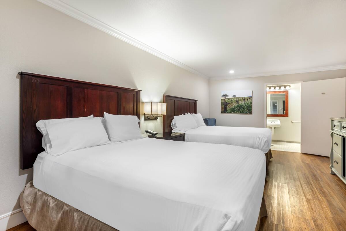 A hotel room with two beds, a nightstand, a lamp, a mirror, a sink, and a cabinet.