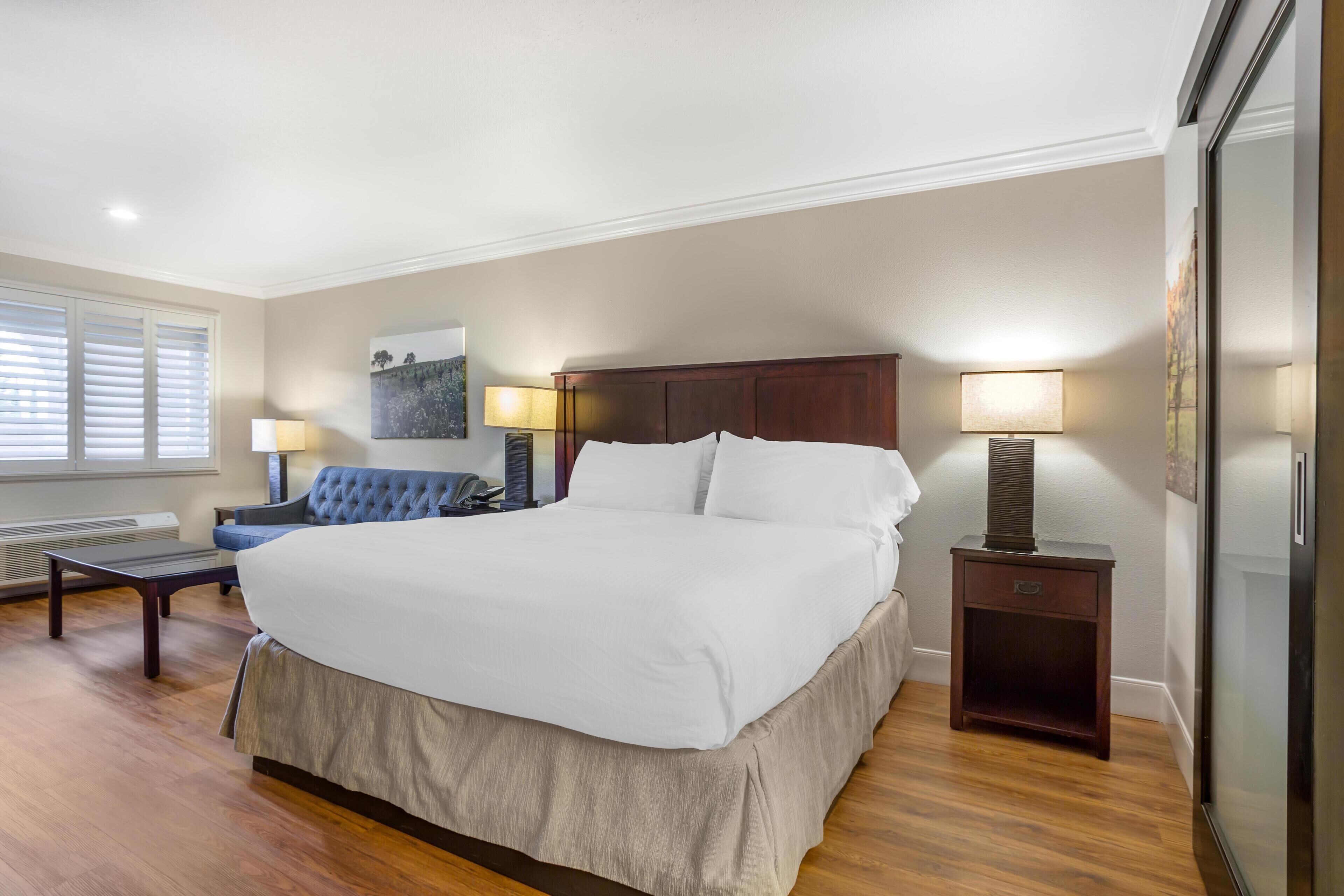 A spacious bedroom with a large bed, a blue sofa, two bedside tables with lamps, a heater, and a large window with blinds.