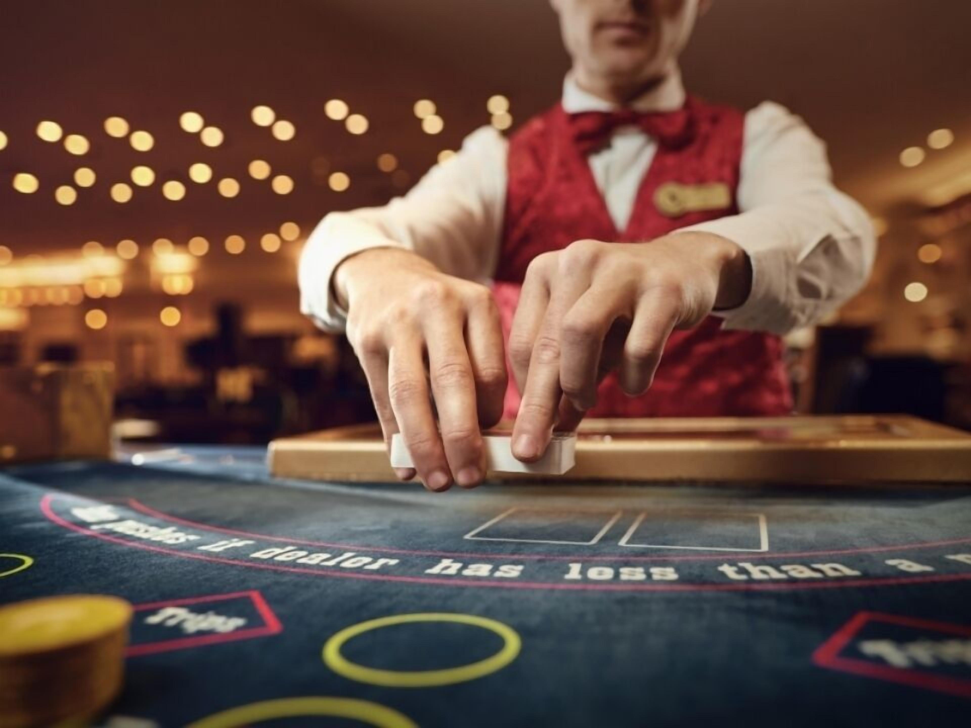 A person dealing cards at a casino table with a roulette wheel visible in the background.