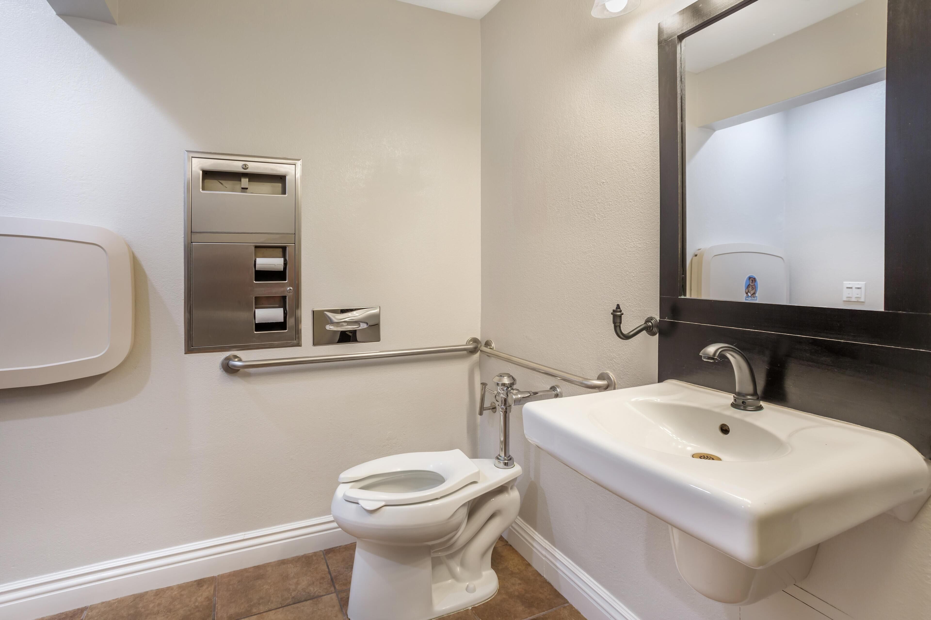 A bathroom with a white toilet, a sink, and a mirror. There is a handrail and a paper towel dispenser.