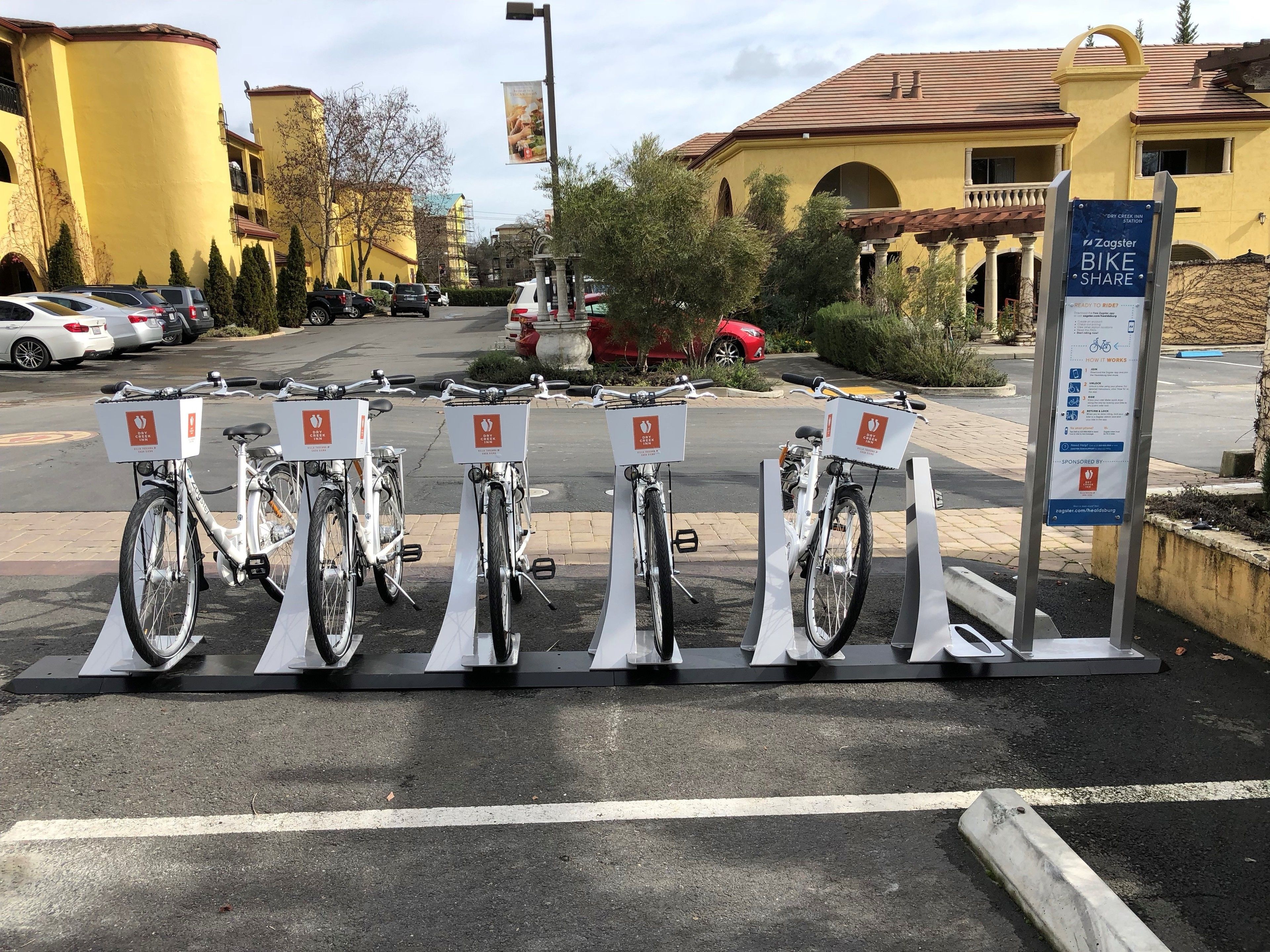 A bike-sharing station with five bicycles, a parking lot, and a building with a yellow facade in the background.