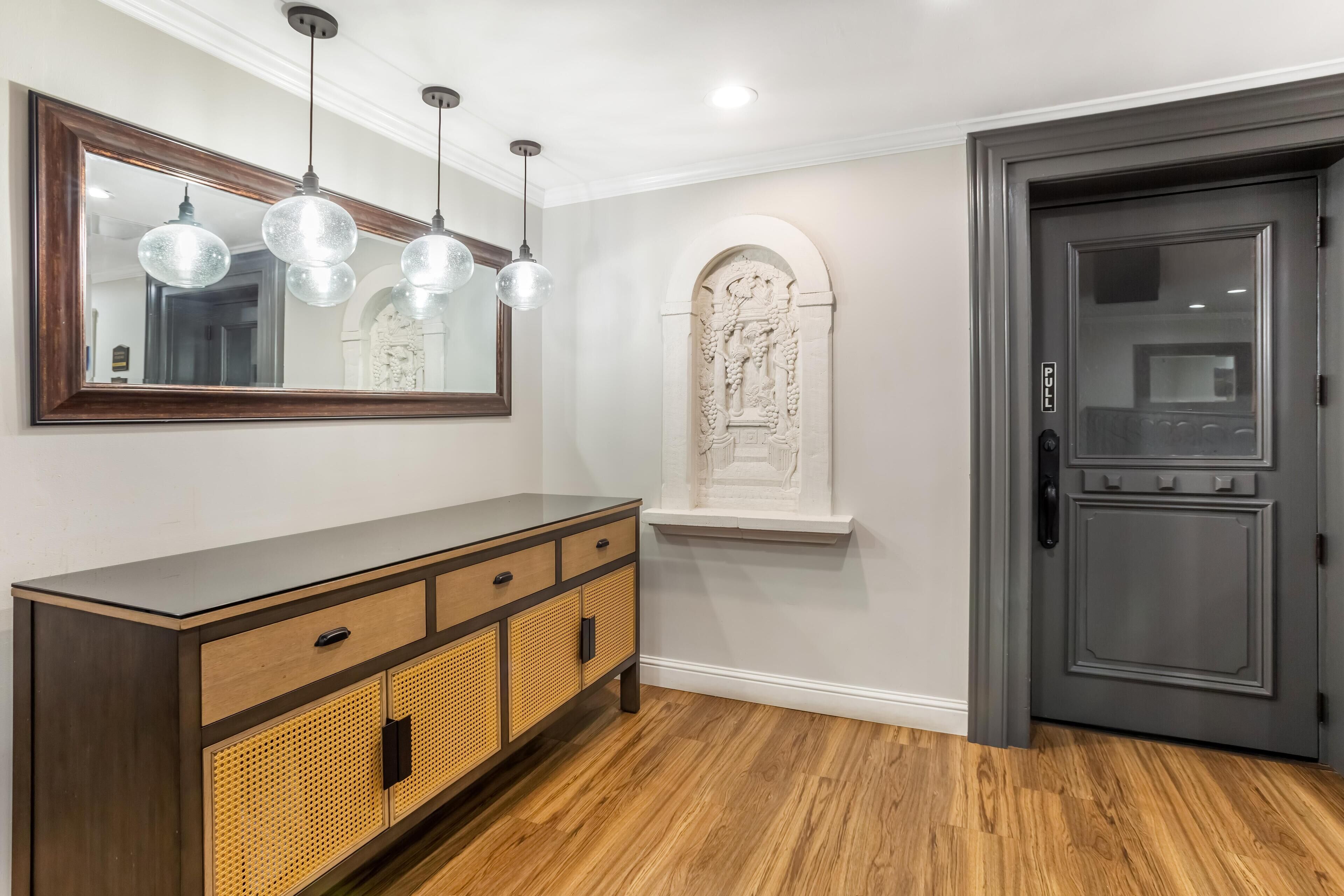 Un pasillo con suelo de madera, un mueble oscuro con cajones, un espejo grande y una puerta con panel de vidrio satinado.