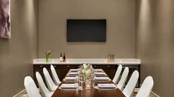 A conference room with a long table, white chairs, a flat-screen TV mounted on the wall, and a small side table with a vase of flowers and candles.