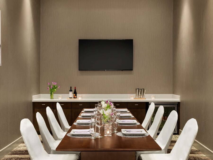 A conference room with a long table, white chairs, a flat-screen TV mounted on the wall, and a small side table with a vase of flowers and candles.