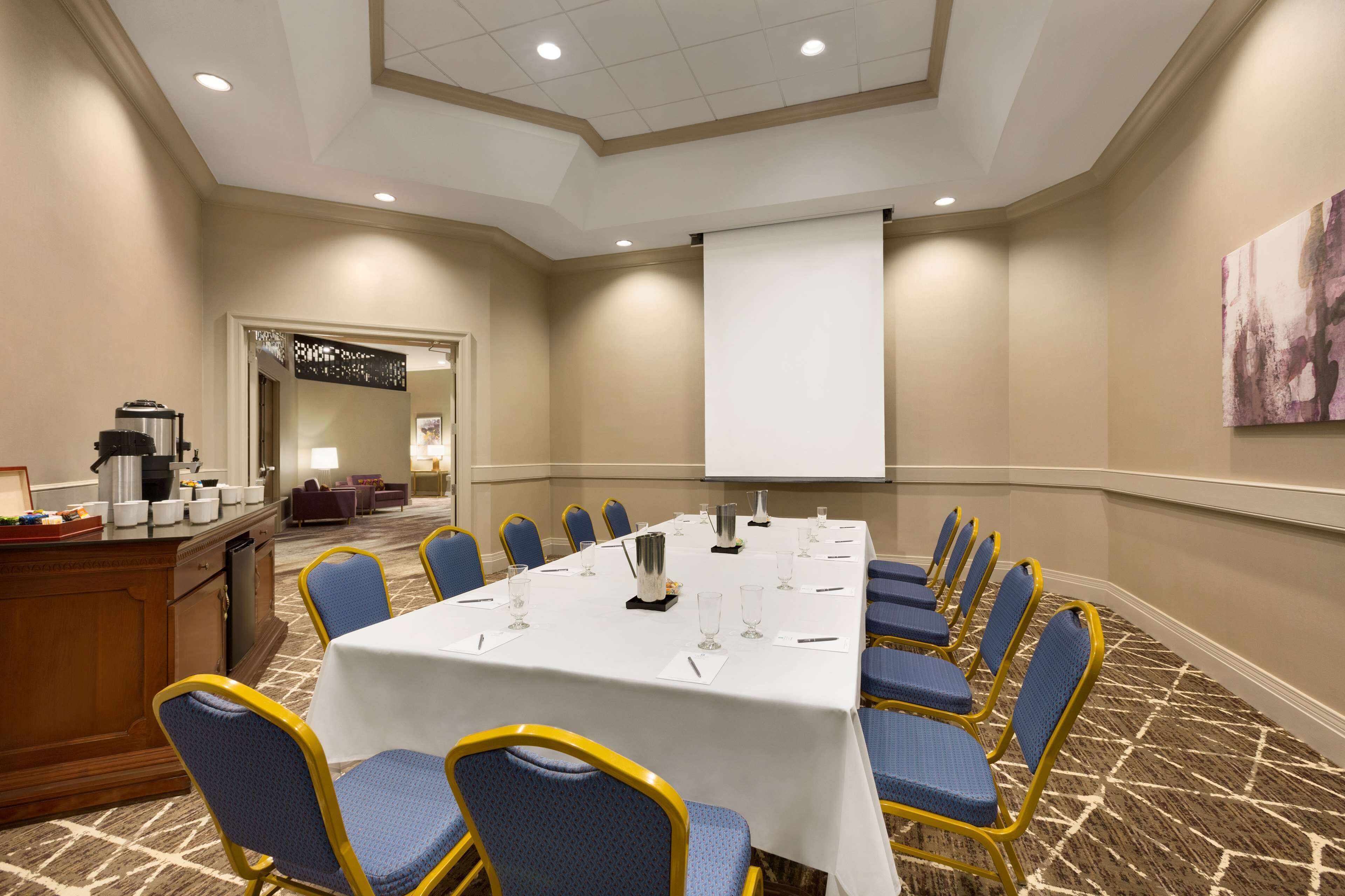A conference room with a long table, blue chairs, a projector screen, and a coffee station.