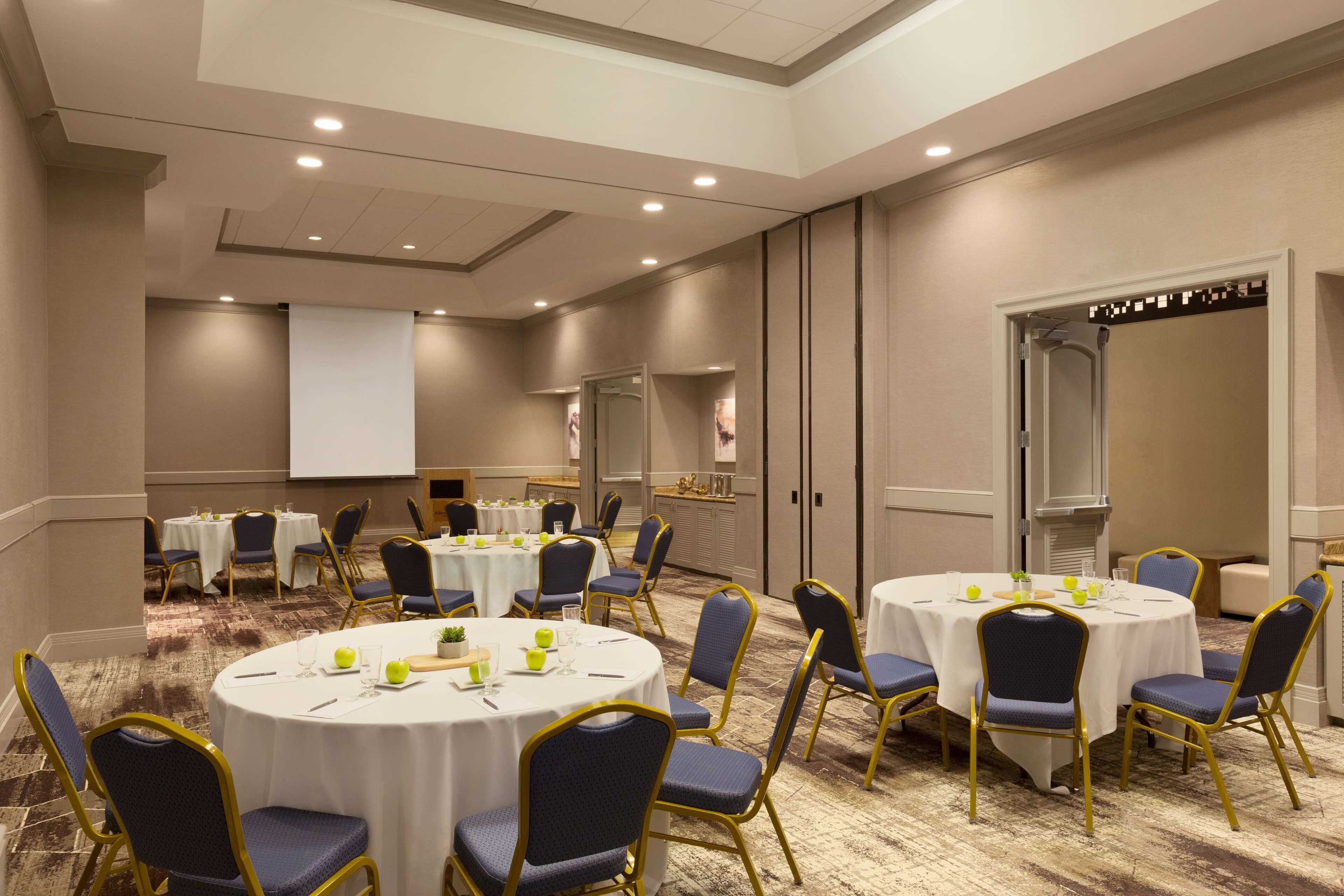 Un salón de conferencias con mesas redondas, sillas y una pantalla para proyector.
