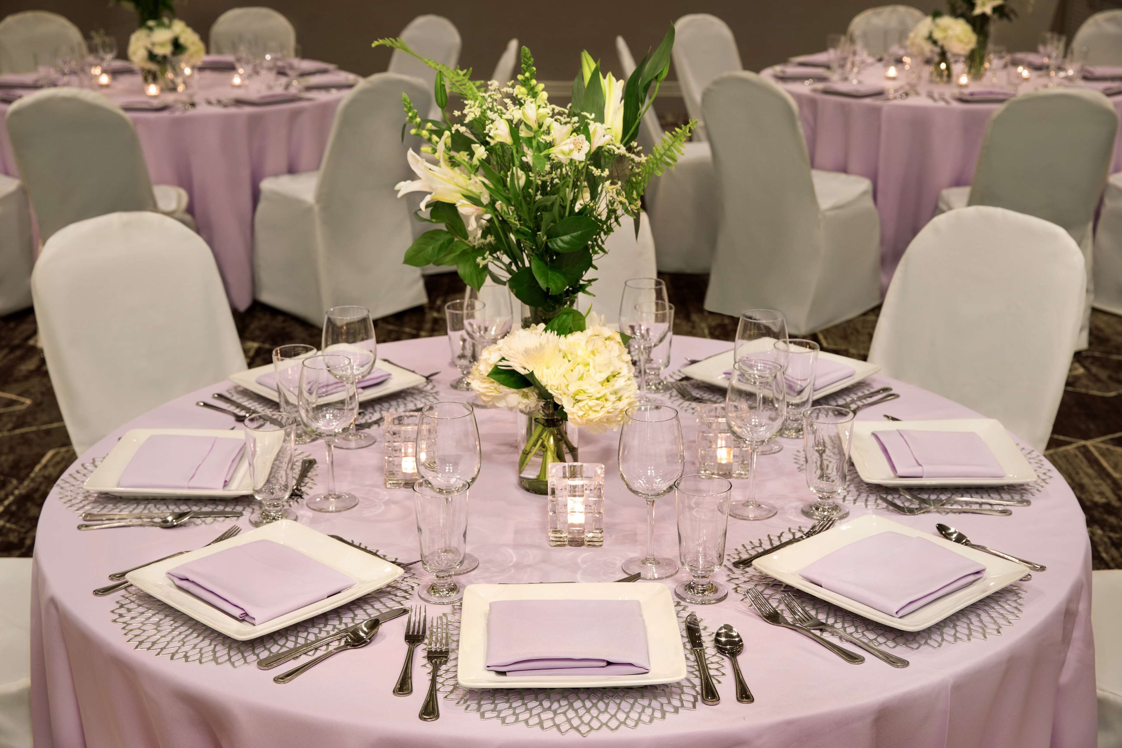 A round table set with white plates, silverware, and clear glasses, surrounded by white chairs.