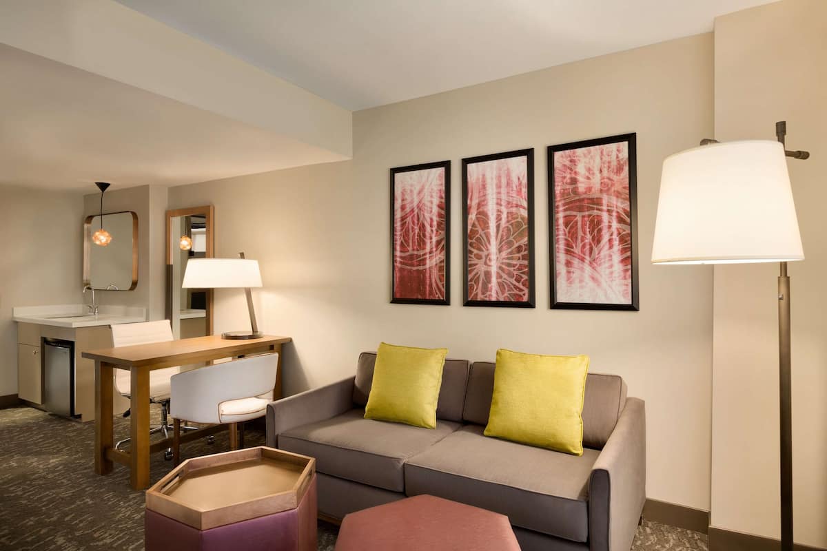 Ein Hotelzimmer mit einer Couch, einem kleinen Tisch, einem Sessel, einem Schreibtisch, einer Lampe und Kunstwerken an der Wand.