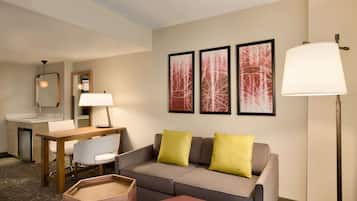 A hotel room with a sofa, a small table, a chair, a desk, a lamp, and artwork on the wall.