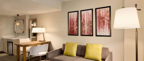 A hotel room with a sofa, a small table, a chair, a desk, a lamp, and artwork on the wall.