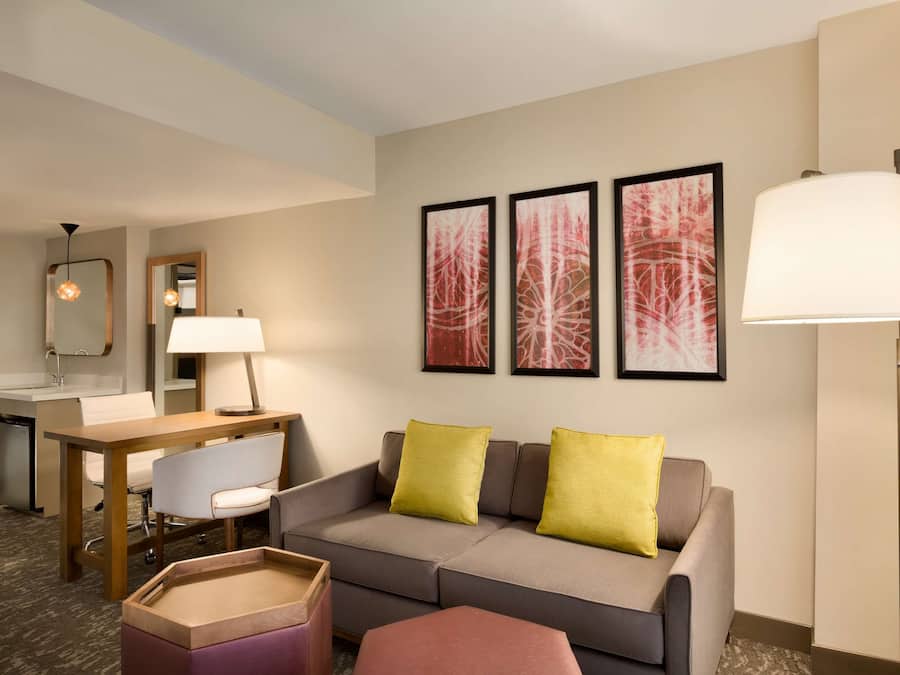 A hotel room with a sofa, a small table, a chair, a desk, a lamp, and artwork on the wall.
