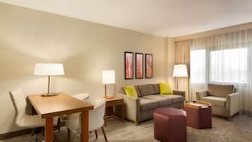 Une chambre d’hôtel avec un bureau en bois, deux chaises, un canapé avec des coussins, un petit pouf et une fenêtre avec des rideaux.
