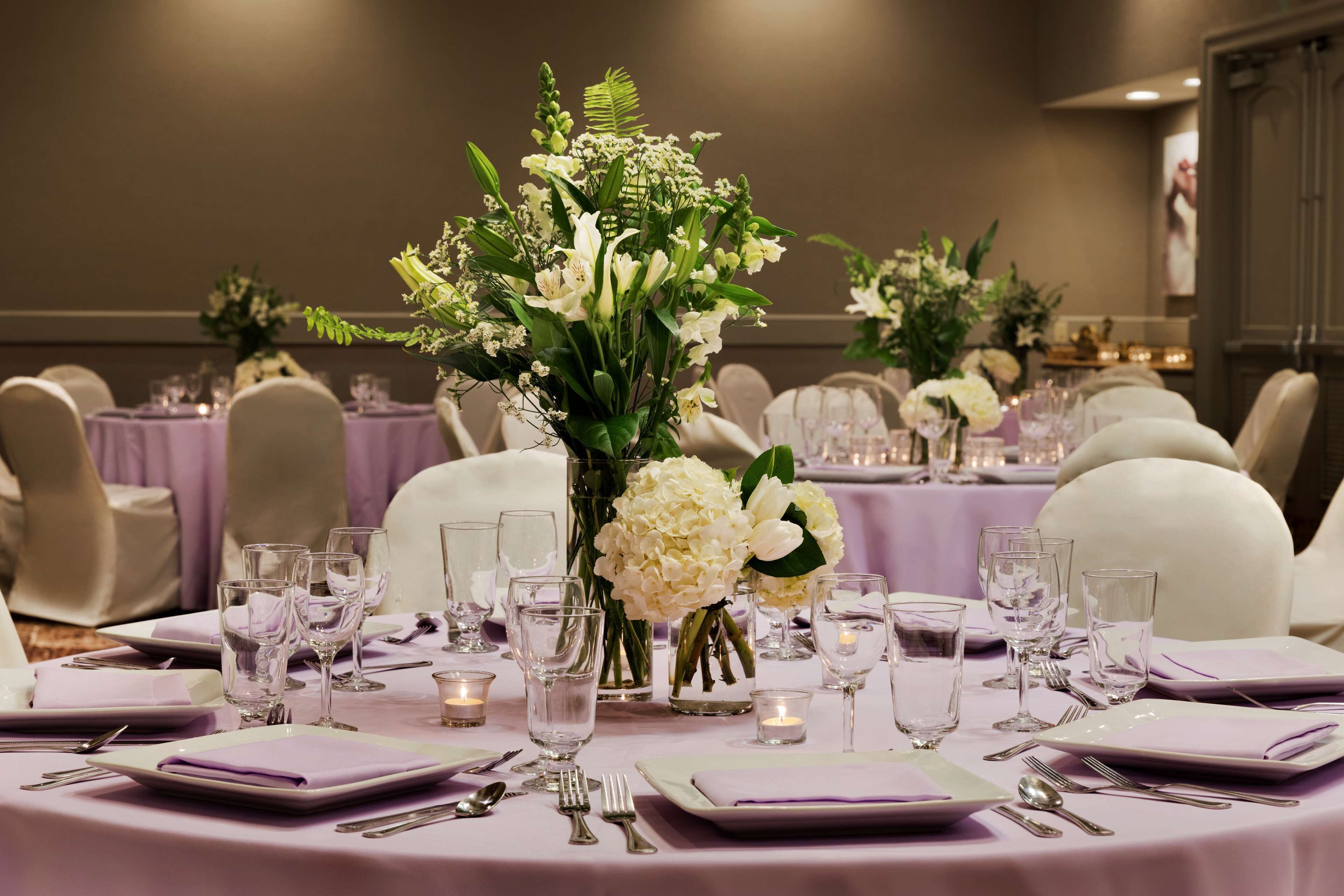 Un salón de banquetes con mesas redondas dispuestas para un evento formal, con centros de mesa florales y velas.