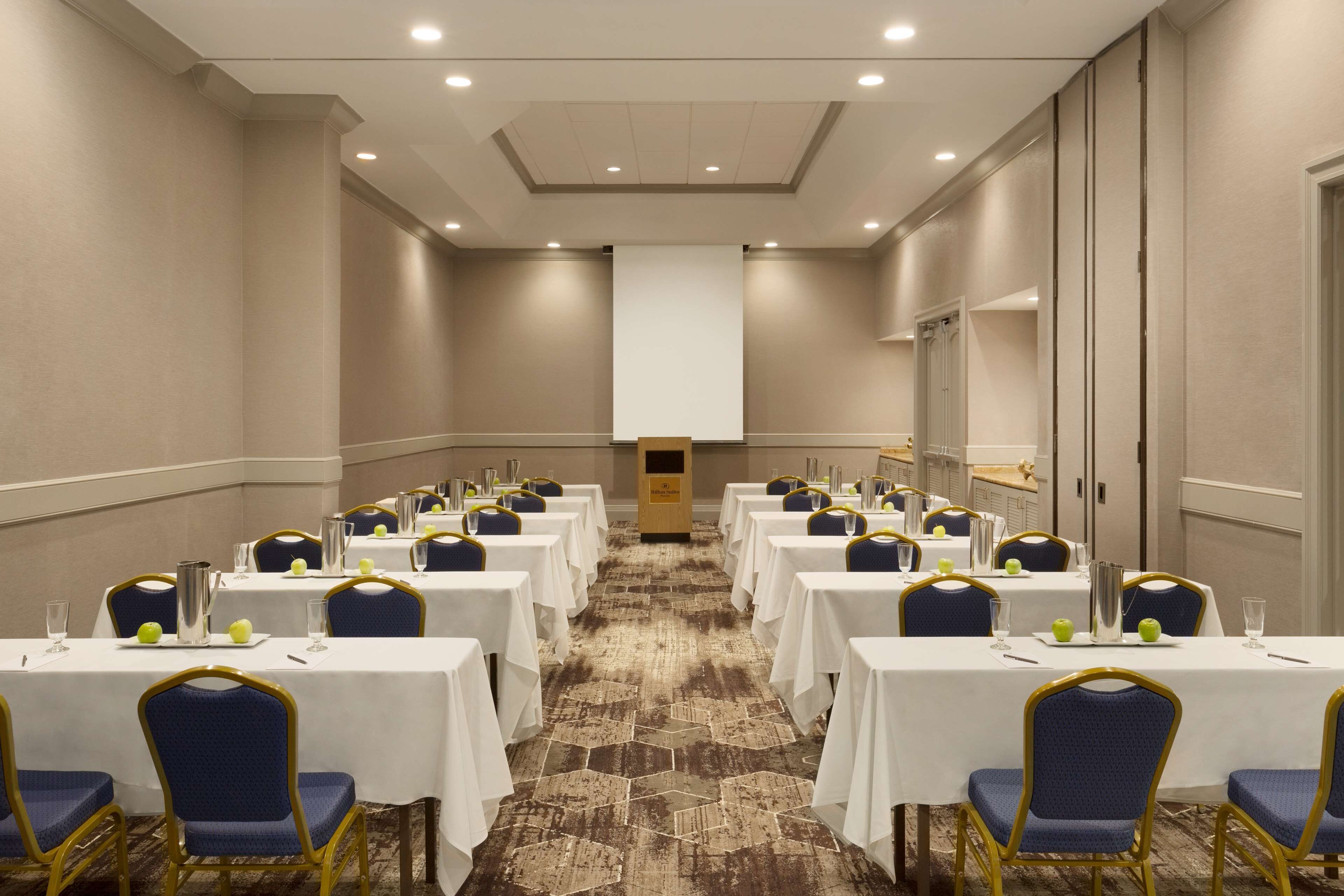 Un salón de conferencias con filas de mesas y sillas, un atril y piso alfombrado.