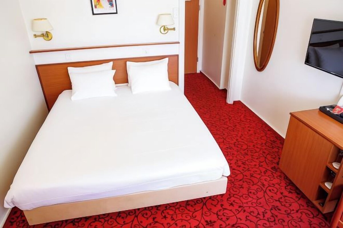 Een hotelkamer met een net opgemaakt bed, een houten hoofdbord, een rood karpet met patroon, een klein houten nachtkastje, en een televisie aan de muur.