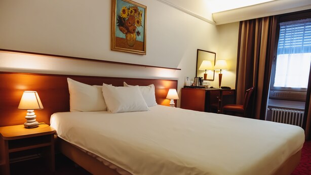Habitación de hotel con una cama grande, dos mesitas de noche con lámparas, un escritorio con una silla, un cuadro en la pared y una ventana con cortinas.