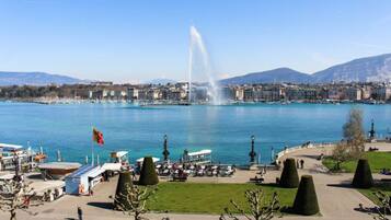 Un paisaje urbano frente al agua, con una fuente destacada, un puerto deportivo con barcos y una vista despejada de montañas distantes.
