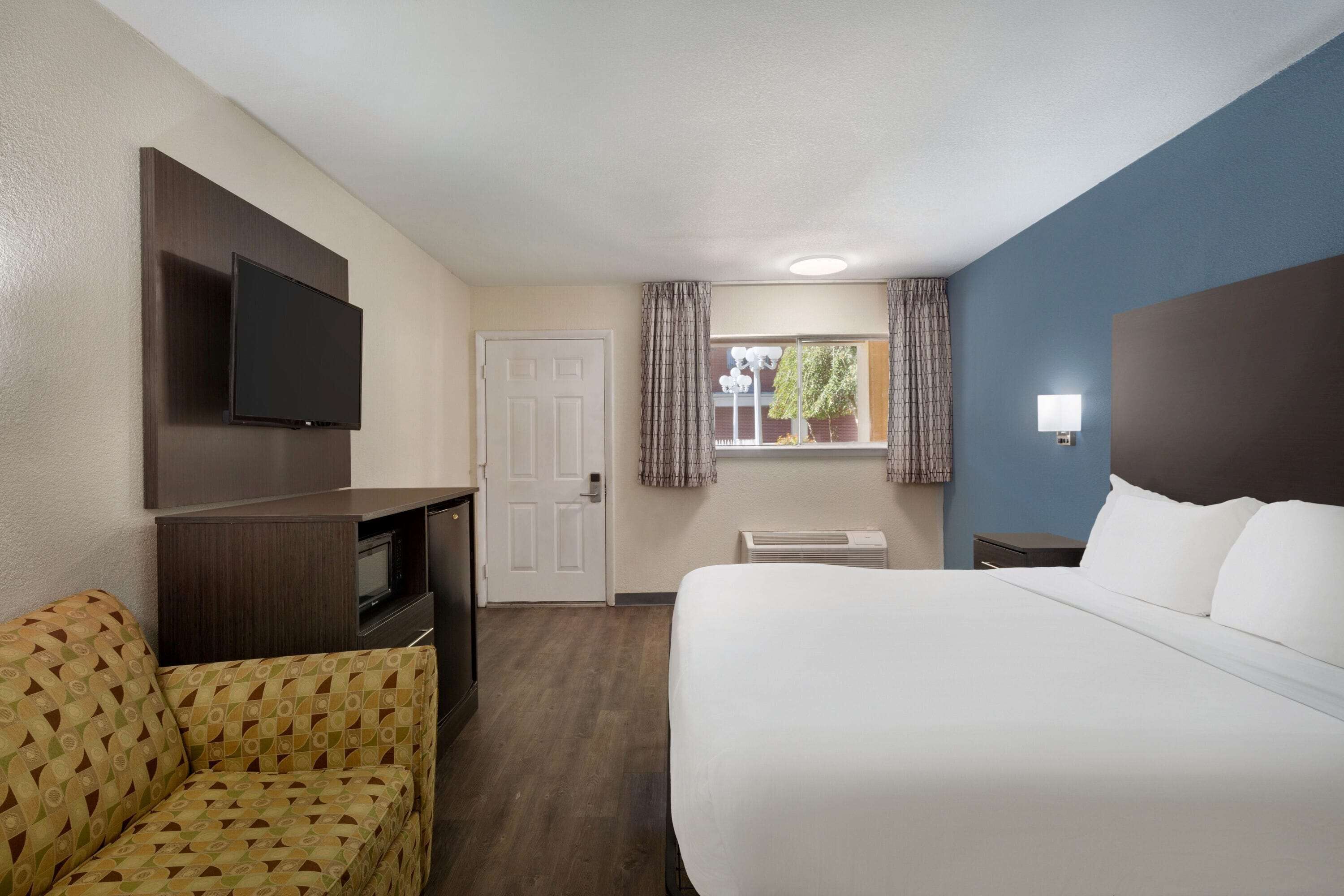 A hotel room with a bed, a sofa, a TV mounted on the wall, a window with curtains, and a bedside table.
