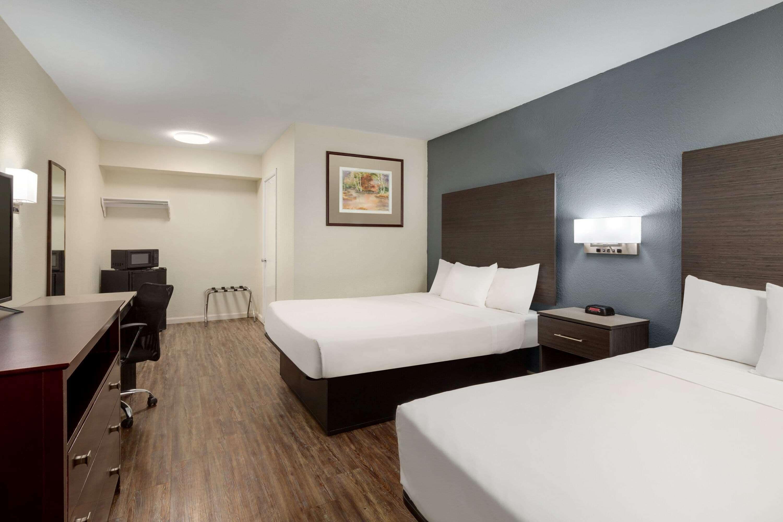 A hotel room with two beds, a desk, a chair, a small table, a nightstand, a lamp, a picture on the wall, and a ceiling light.