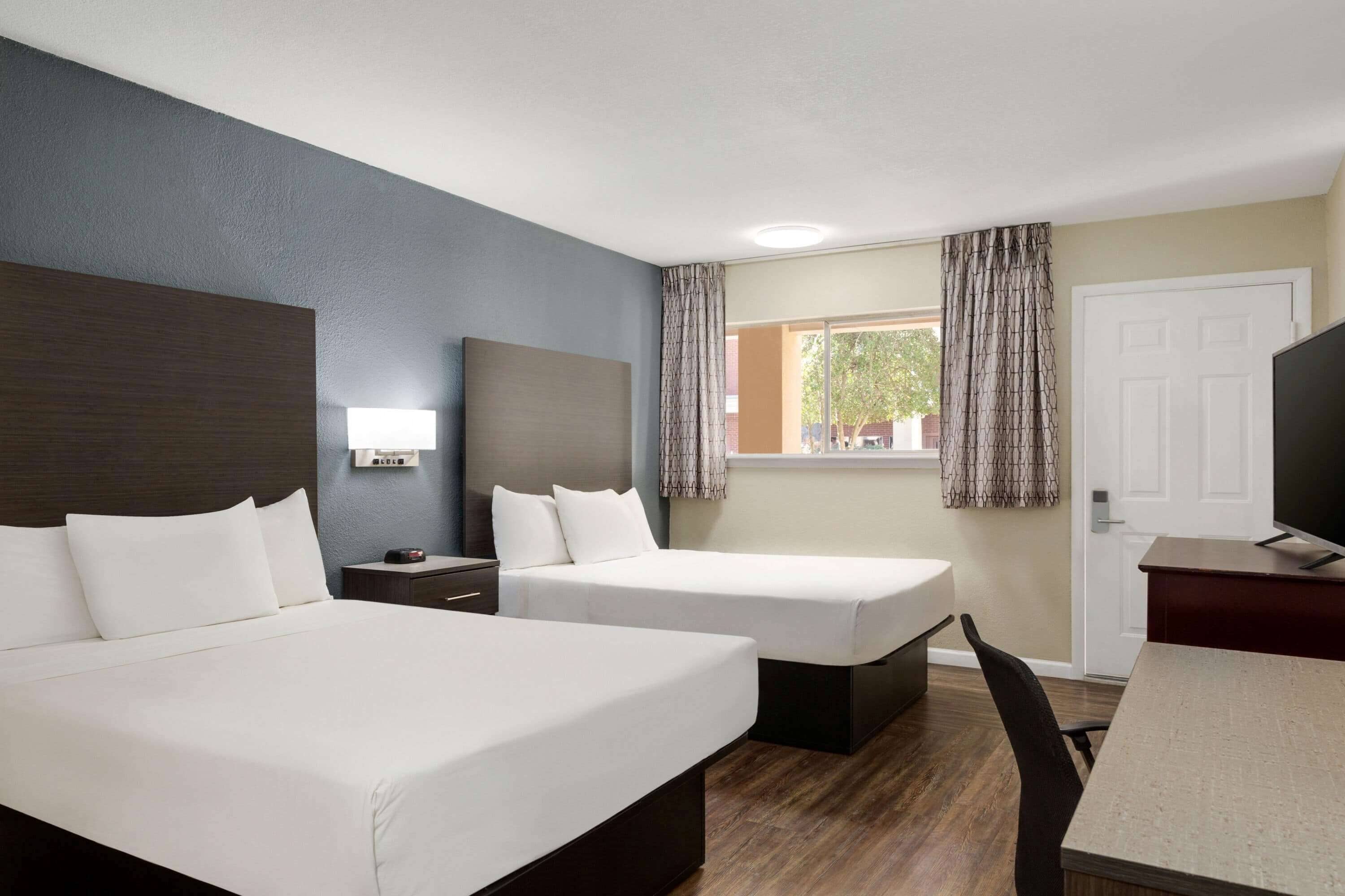 Habitación de hotel con dos camas, un escritorio, una silla, un televisor y una ventana con cortinas.