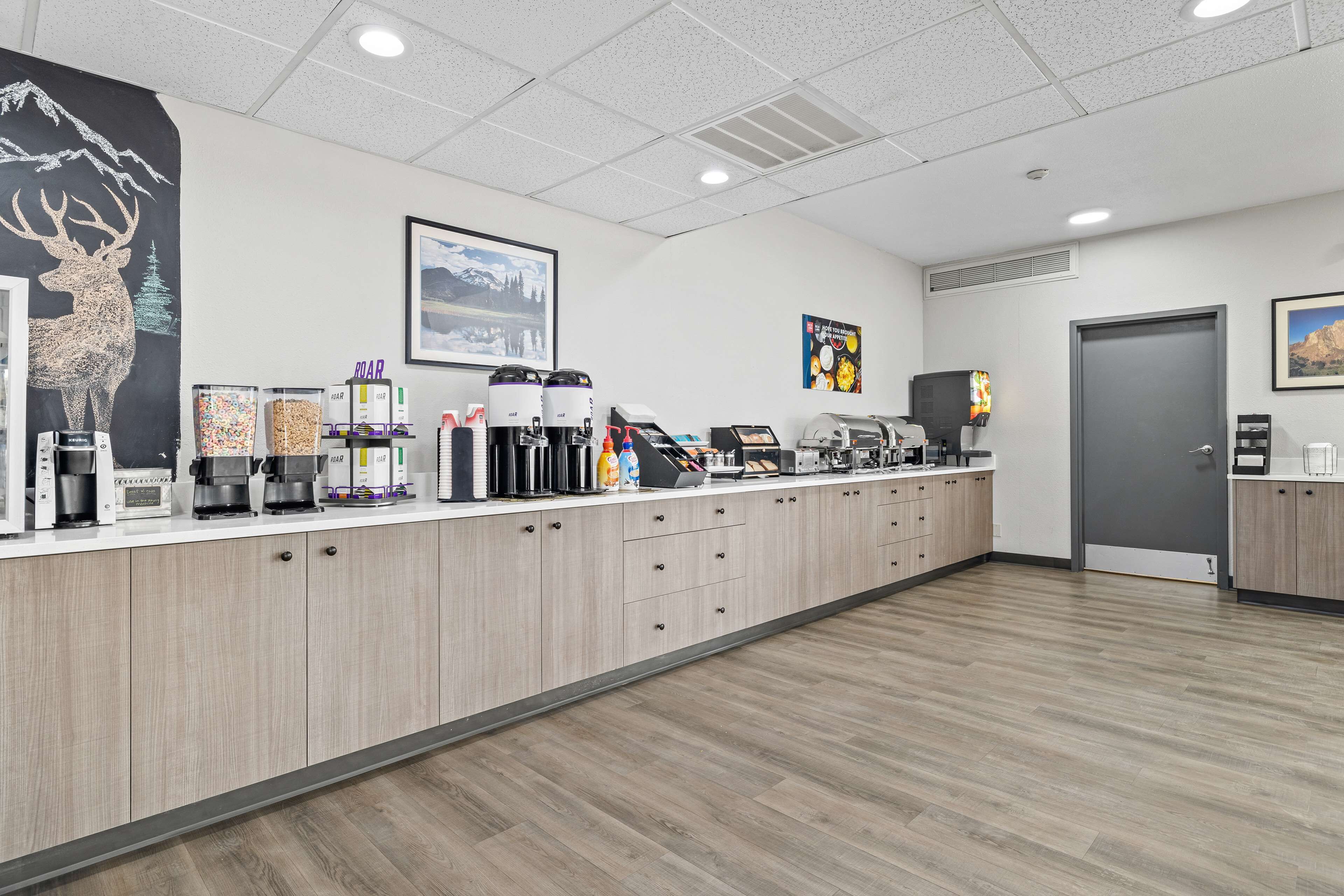 A modern coffee station with multiple coffee machines, a large poster of a deer, and a framed landscape picture on the wall.