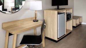Habitación de hotel con escritorio de madera, una silla negra, televisor, espejo y mueble con microondas.